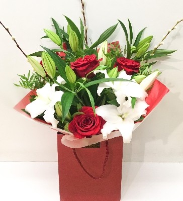 Hand-tied bouquet with red Freedom roses, white lilies, willow accents and foliage, wrapped in luxury cellophane and gift bag.