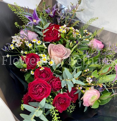Hand-tied bouquet featuring ranunculus, roses, matricaria, clematis, and seasonal florals, presented in signature black wrap with a satin bow.