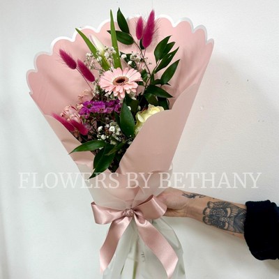 Hand-tied pink bouquet with seasonal florist’s choice florals and greenery, wrapped in scalloped blush pink paper with a satin ribbon.