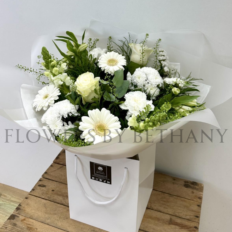 White and green hand-tied bouquet with mixed seasonal florals and foliage, presented in a white gift bag.