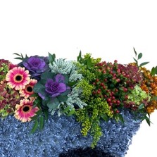 Wheelbarrow-shaped funeral tribute filled with vibrant seasonal flowers, with the option to include fresh fruit and vegetables, measuring approximately 91cm x 57cm.