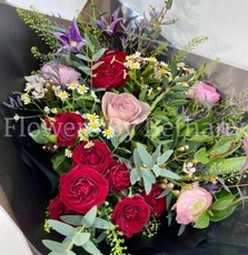 Hand-tied bouquet featuring ranunculus, roses, matricaria, clematis, and seasonal florals, presented in signature black wrap with a satin bow.