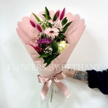 Hand-tied pink bouquet with seasonal florist’s choice florals and greenery, wrapped in scalloped blush pink paper with a satin ribbon.