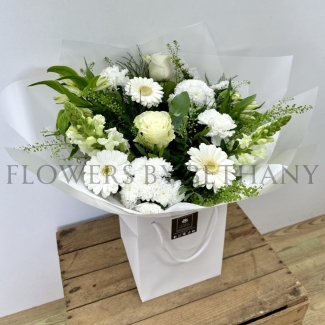 White and green hand-tied bouquet with mixed seasonal florals and foliage, presented in a white gift bag.